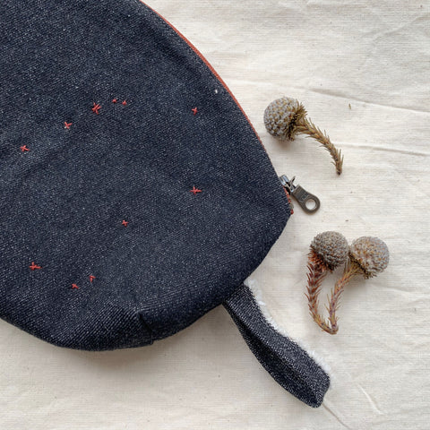 showing the outside of the purse which is a dark blue denim. it is only showing one half of the pouch and also shows a loop to hang the pouch. there are three small seed pods on the ivory linen as decoration