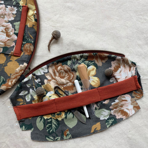 a picture of the purse before it has been sewed together. it shows one side with a rib band across it divided into three parts. the middle section holds some sewing snips, seam unpicked and awl. the inside of the pouch is a grey floral needle cord.