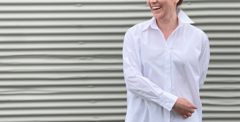 a white girl standing against a grey corrugated iron backdrop she wears a white shirt and is laughing