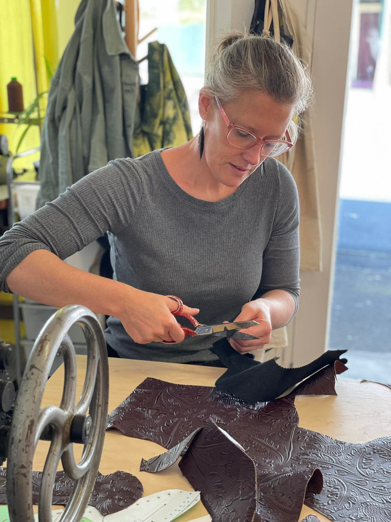 a white lady cutting out leather with scissors while sat a wooden bench