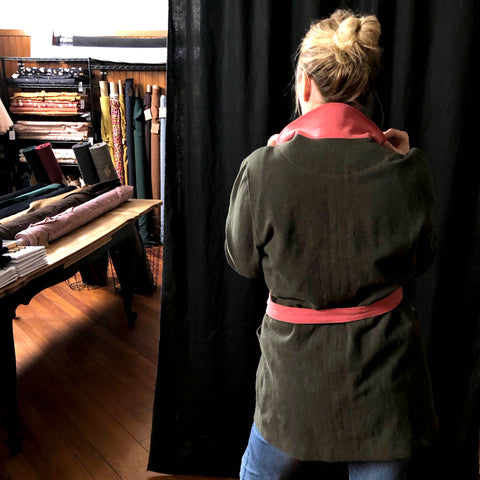a picture of the coat on the lady. this time the photo is from the back. in the background of the photo there are bolts of coloured fabric on shelves and on a table. 