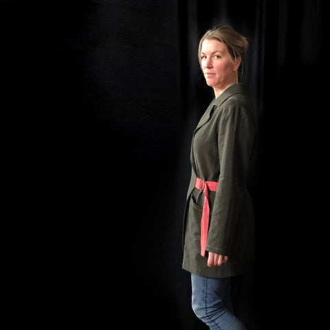 a picture of a lady looking moodily at the camera, it may have been taken at the end of the shoot when she was a bit over it. It is a side view of the lady and it is a dark background. She is wearing a khaki green coat made of linen with a pink belt. you can see a portion of her legs in jeans to show the coat is thigh length.