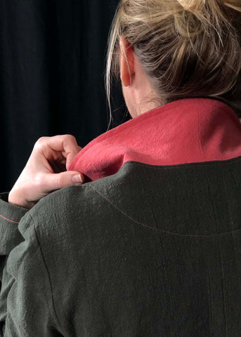 a close up of the lady from the earlier photos but taken from the back. It shows  the collar of the Sienna Maker Jacket up and the lady's hand is holding the collar. The back of the lady's head is showing and her hair is pulled up in a messy bun. The photo shows the pink topstitching on the green jacket.