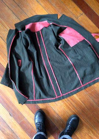 a photo of the inside of the jacket. It is laid on a wooden floor and is opened out. It shows how all of the seams have been bound by bias binding. It also shows the pink interior pocket with the jacket facing slightly over the top of the pocket.