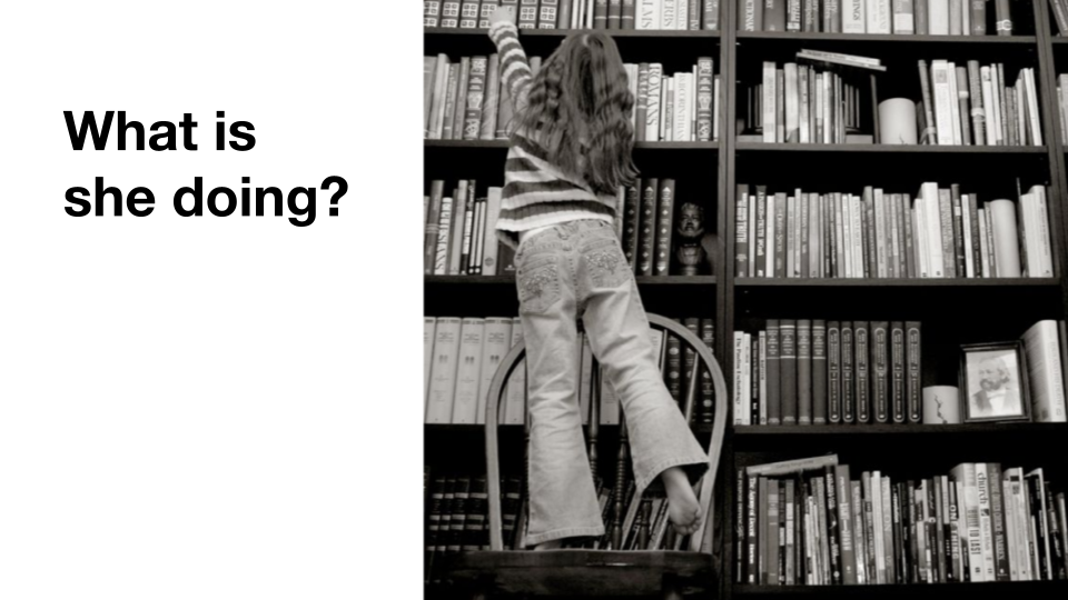 A girl standing on a chair reaching for a book.