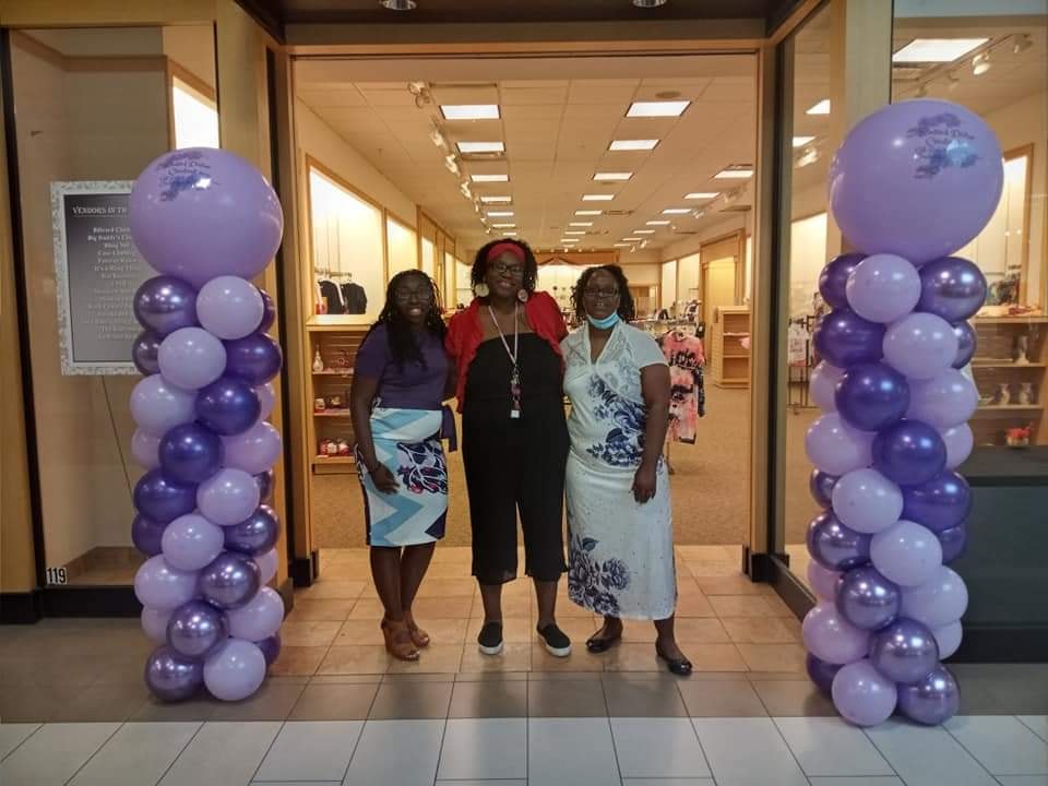 Three women standing in front of a storefront surrounded by purple balloons.