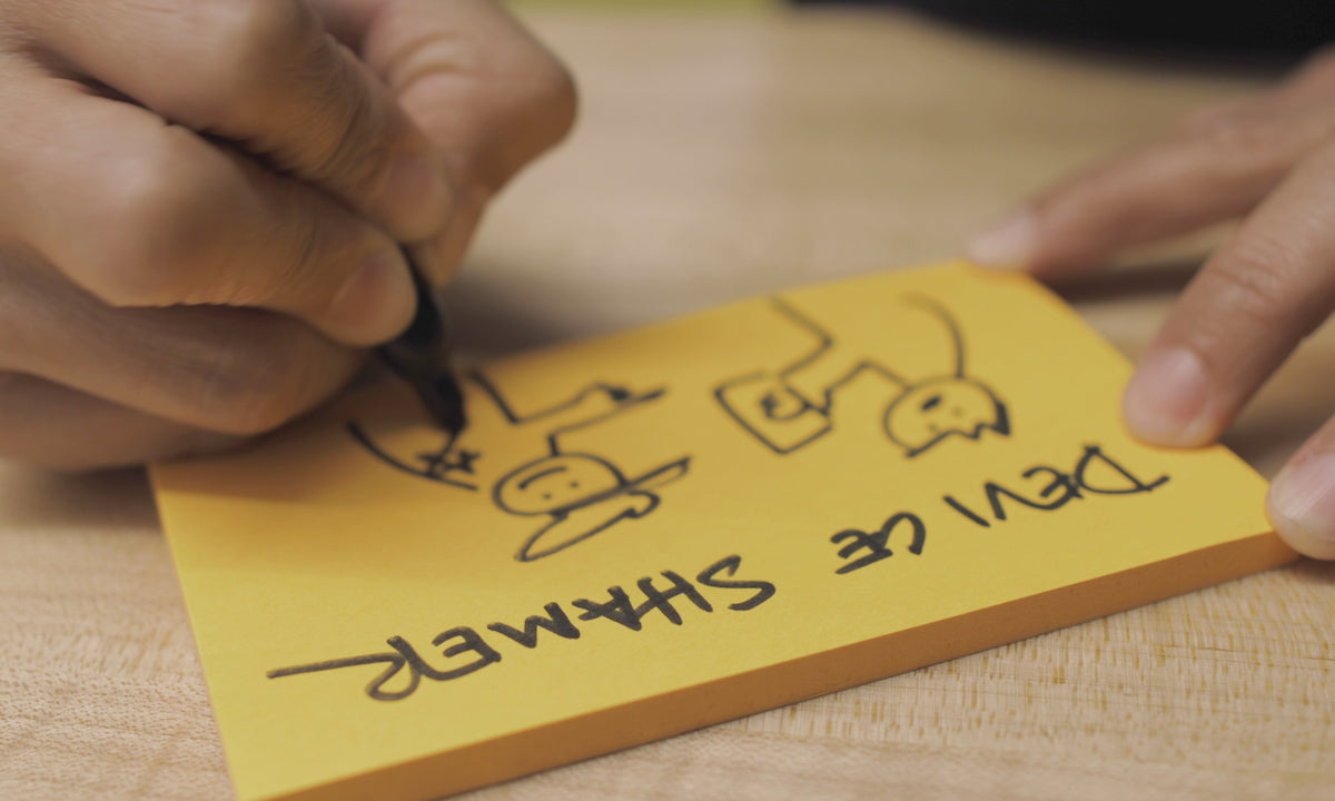 A person draws two stick figures with a sharpie on a yellow sticky note.