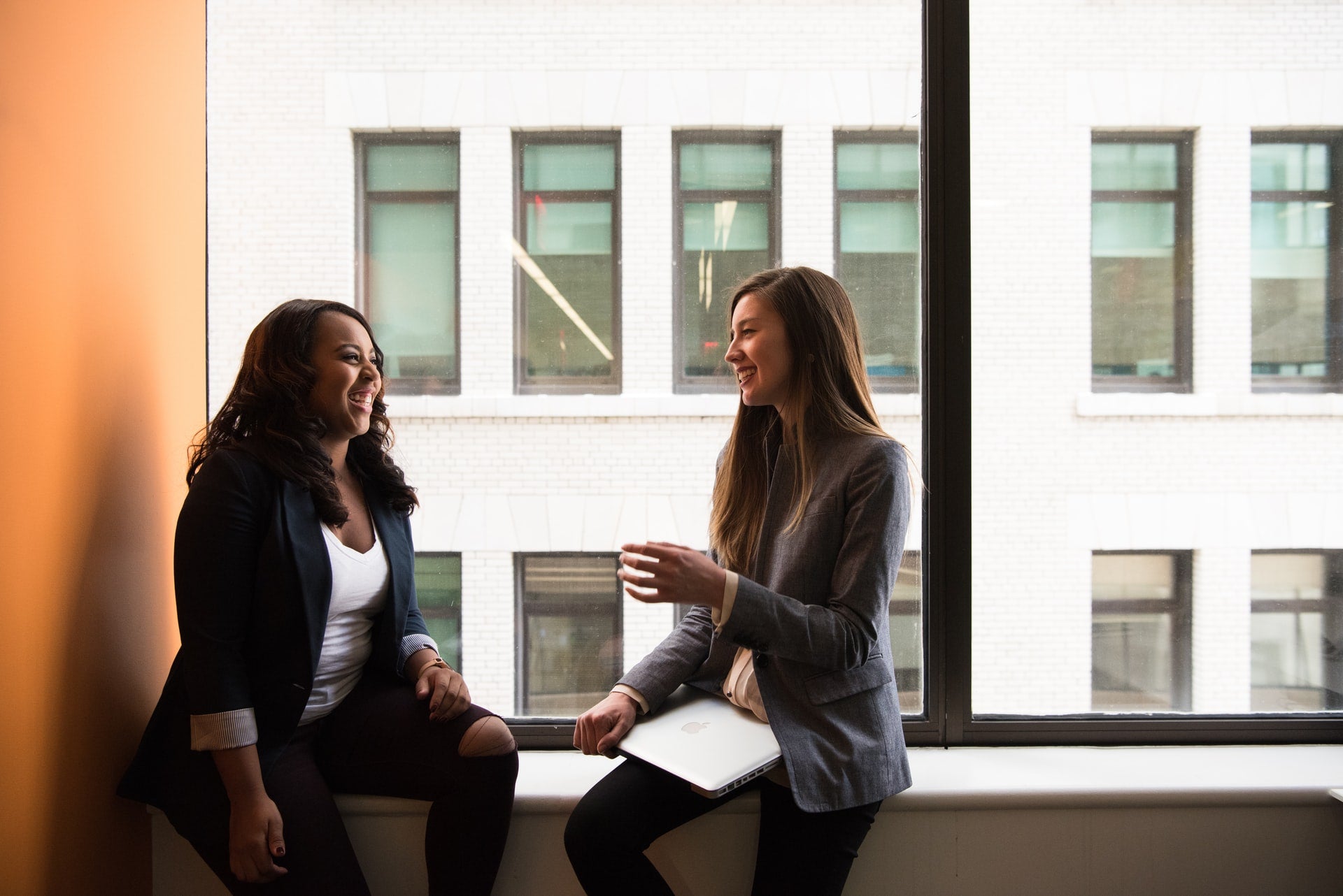 Two women sitting in front of a window smiling at each other.