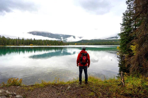 A person in a red jacket with a red waterproof backpack standing on the shore of a lake.