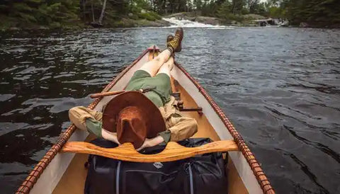 A person laying with their head on black waterproof duffel bag in a wooden canoe on the water.