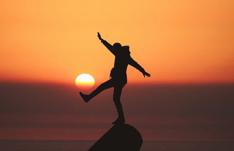 Man hanging off a rock near water, in front of sunset