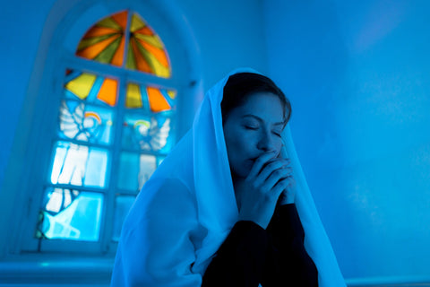 Praying in blue light with a white shawl overhear head in front of a stained glass window.