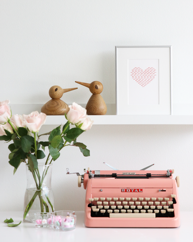 two wooden birds on a shelf next to a framed heart. Image is styled wiht pink roses and a pink typewriter