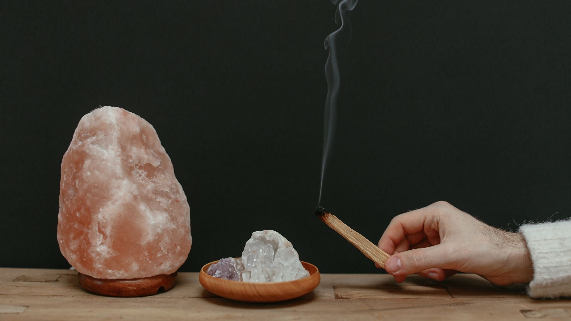 A lit stick of Palo Santo burning next to a salt lamp and crystal on a dish