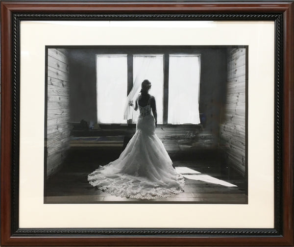 Dramatic grayscale photograph of bride in lacy wedding dress looking out a window custom framed in traditional walnut frame.