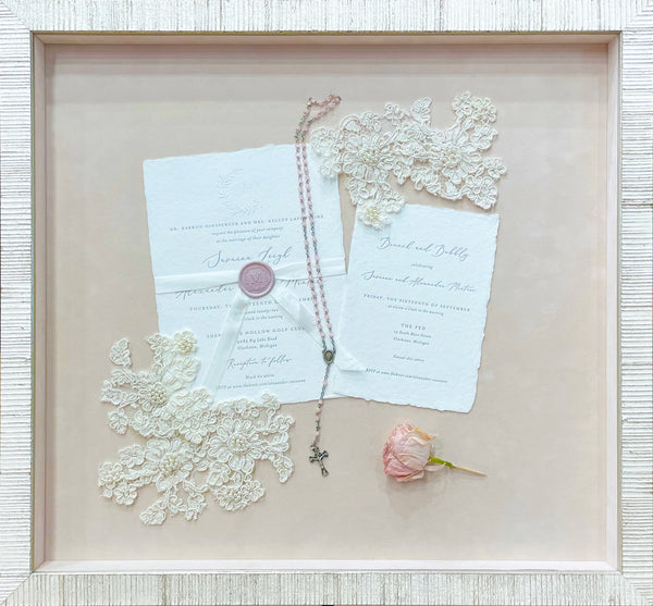 Wedding artifacts, including lace from the wedding gown, invitations, rosary and dried bloom arranged in a luxurious shadowbox with pink suede mat and shimmery white frame.