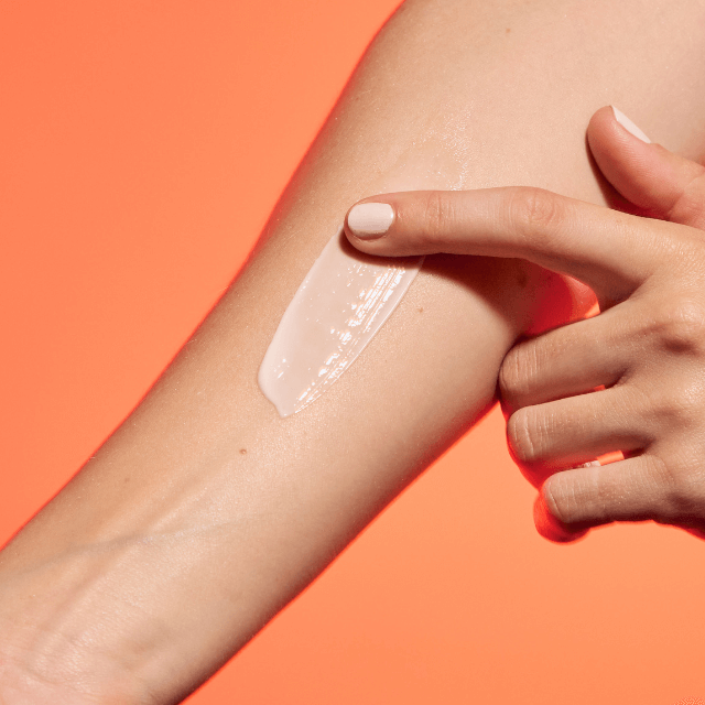 A person applying lotion to their arm against a bright orange background.