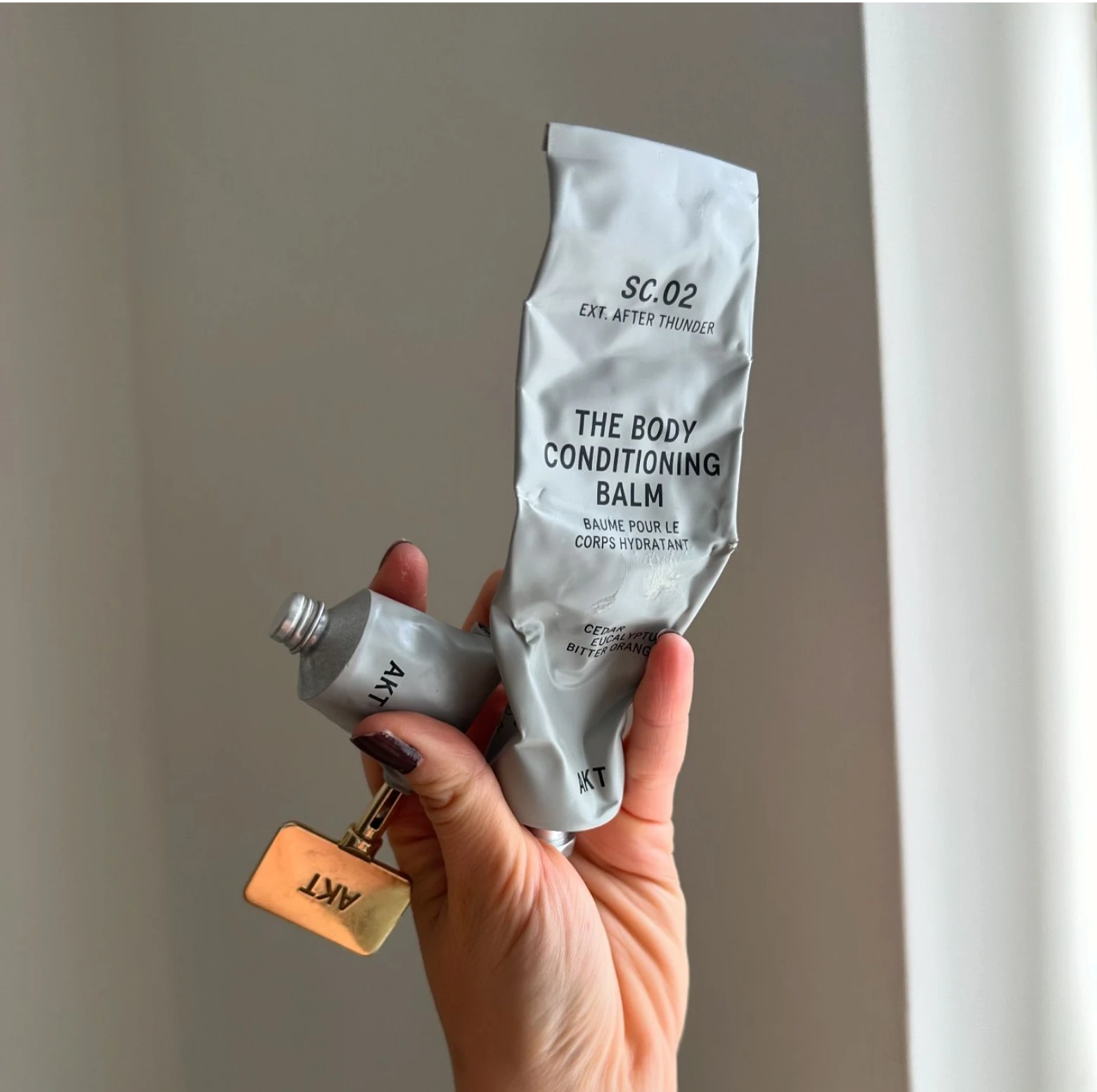 Hand holding a crumpled tube of body conditioning balm near a window.