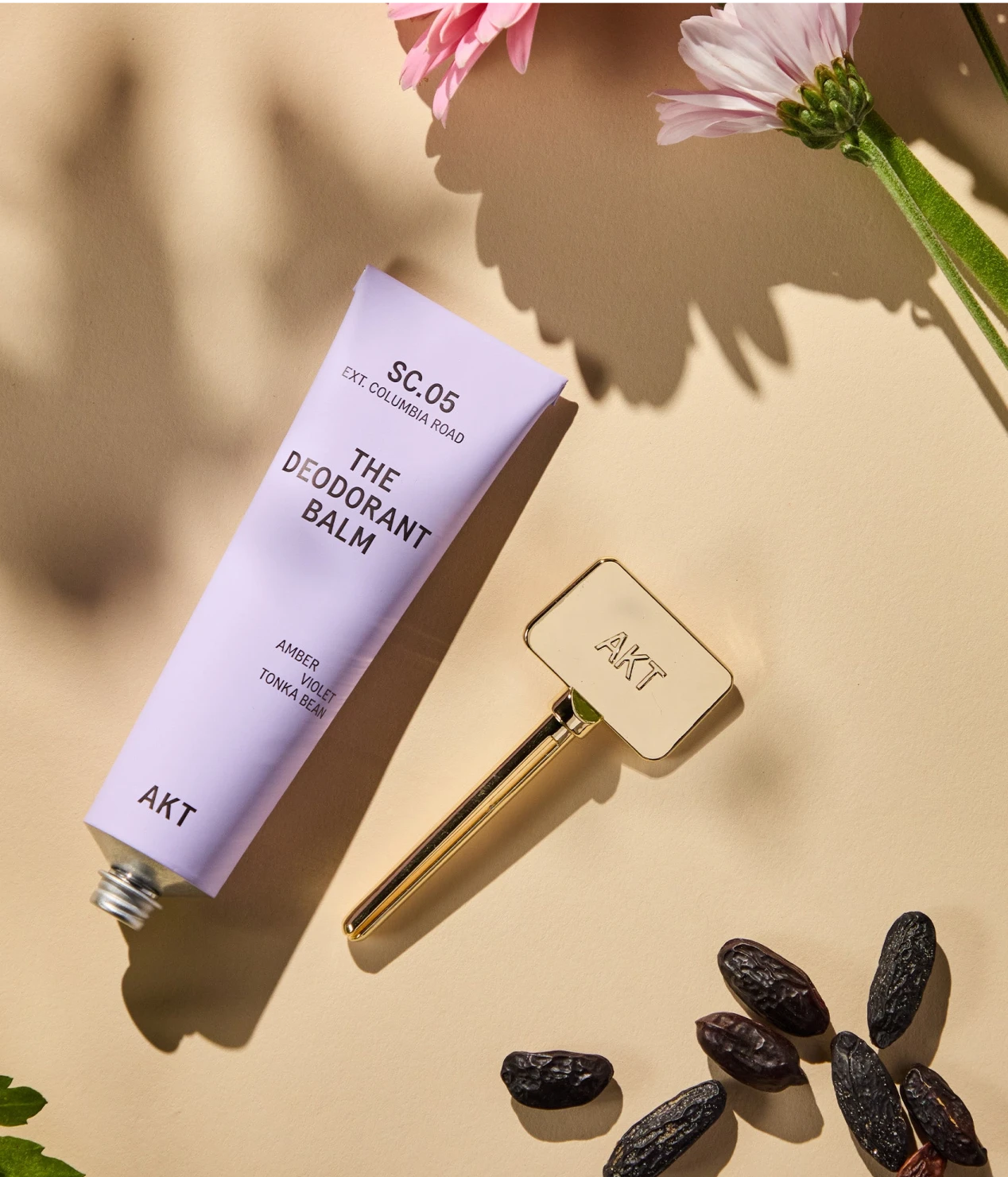 Deodorant balm tube with gold applicator surrounded by flowers and seeds on a beige background.