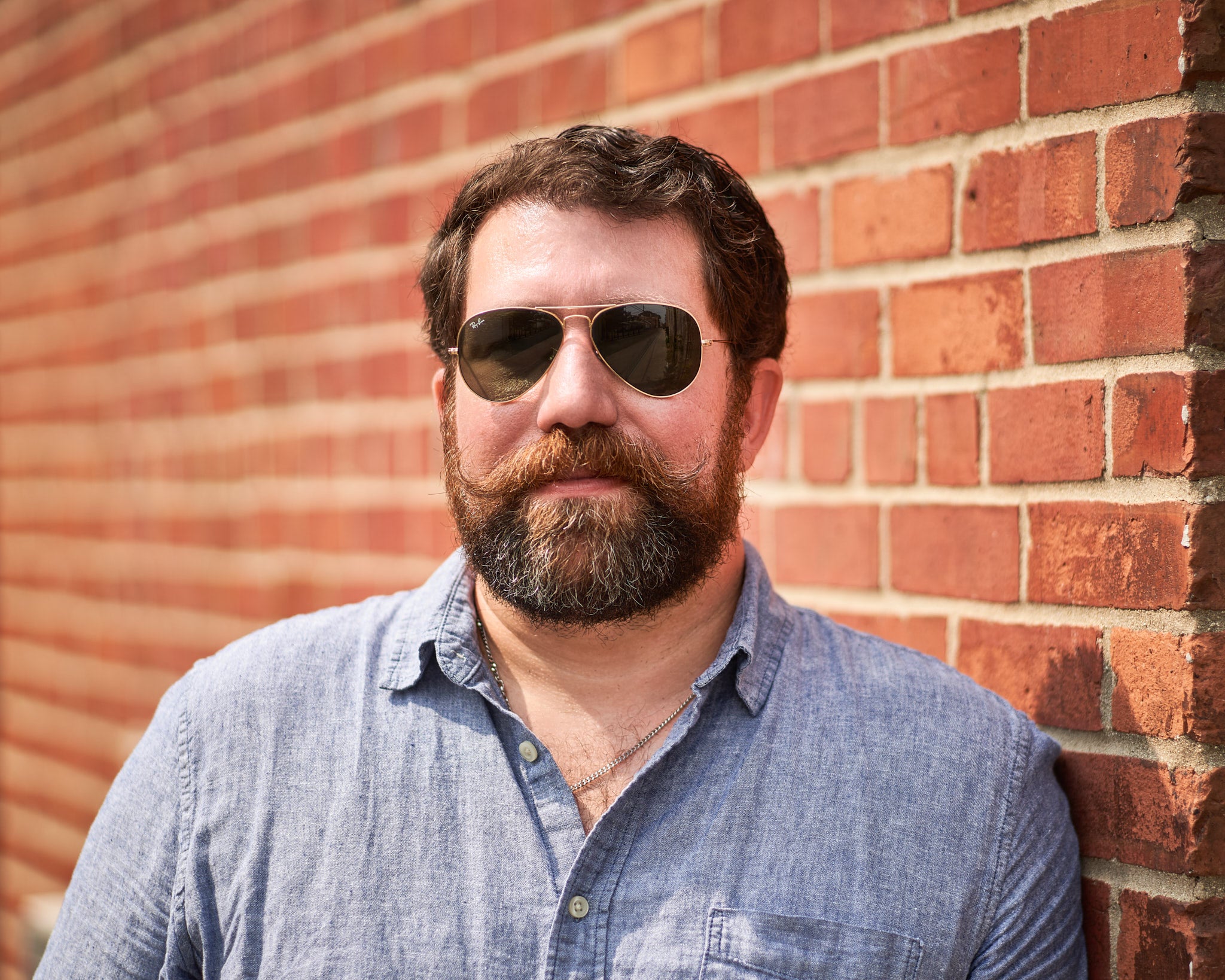 Image of Greg standing in front of brick wall with sunglasses and beard.