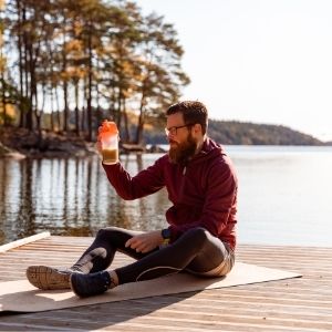 Man sitting on yoga mat on a jetty by the lake looking at a half finished protein shake