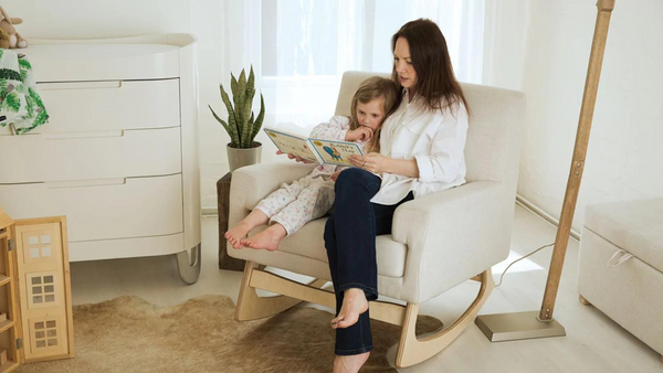 Mutter liest einem Kind ein Buch vor, während sie auf einem Schaukelstuhl sitzt. Gaia Baby Serena Still- und Schaukelstuhl in der Farbe Hafer. Hellgrauer Still- und Schaukelstuhl