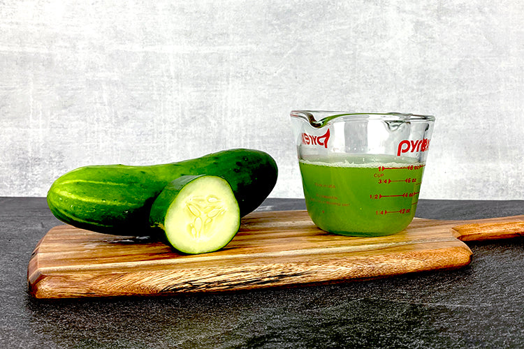 One cucumber and one cucumber slice next to a measuring cup filled with one cup of cucumber juice.