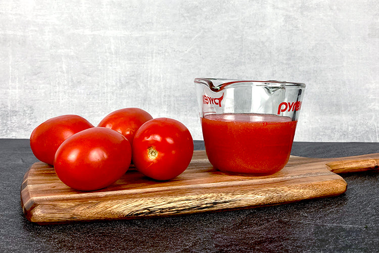 Four tomatoes next to a measuring cup filled with one cup of tomato juice.