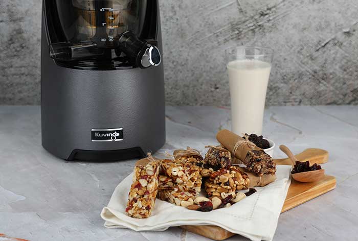 A pile of energy bars in the foreground, with a Kuvings juicer and a glass of milk in the background.