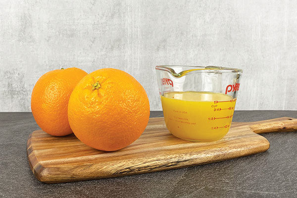 Two oranges next to a measuring cup filled with one cup of orange juice.