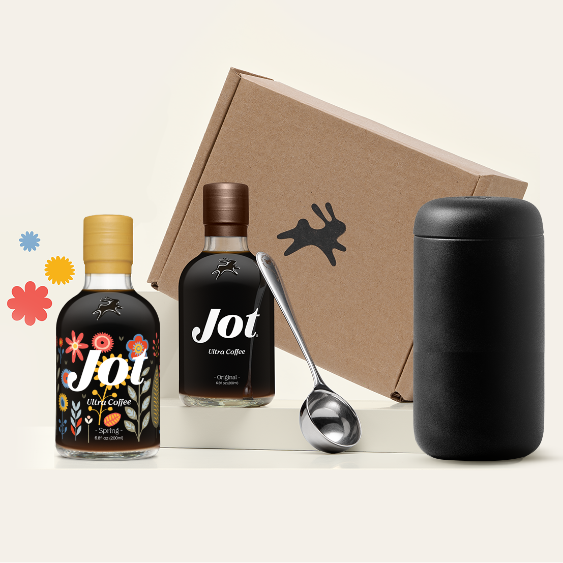 Two bottles of Jot coffee concentrate, a spoon, and a travel mug with cardboard box and decorative flowers.