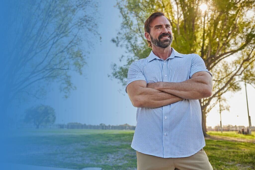 Man with crossed arms standing outdoors near trees on a sunny day.