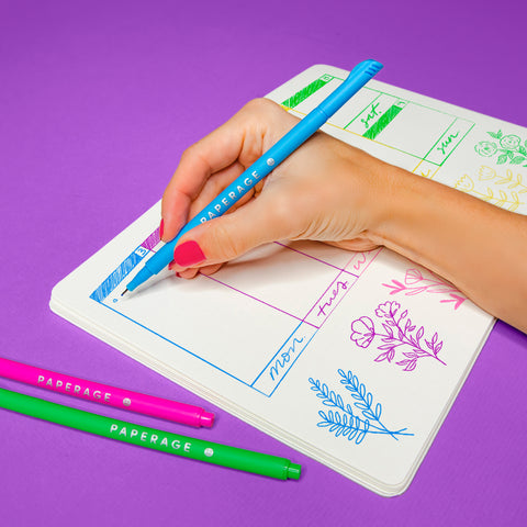 Hand holding blue felt tip pen writing in an open dot journal on a purple background. The pages of the dot journal show a variety of line drawings of flowers, boxes for planning and the days of the week.