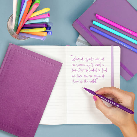 Hand writing a quote in an open journal with pens and books surrounding. The quote in the book reads: "Kindred spirits are not so scarce as I used to think. It's splendid to find out there are so many of them in the world."