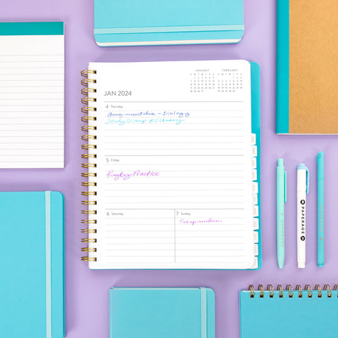 Overhead view of a variety of journals, notebooks, and pens in blue on a light purple background. The center shows on open academic planner with some notes written on the lined pages.