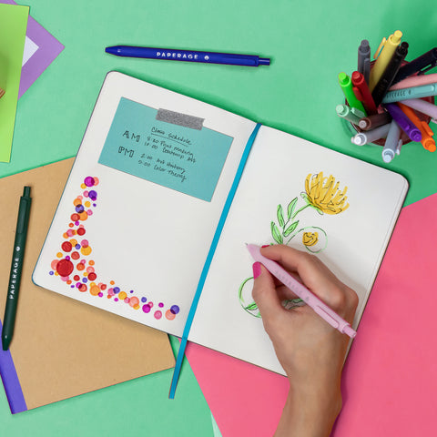 View of an open sketchbook with colorful doodles and a blue card with hand written notes taped to the left page. A hand is drawing a yellow flower with a pink gel pen. A cup of pens and colorful papers are surrounding the open book.