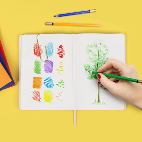 An open sketchbook on a yellow background with 2 colored pencils and a stack of journals. A hand is seen on the right side drawing a tree with a green colored pencil. A variety of colorful scribbles are shown on the left side of the page.