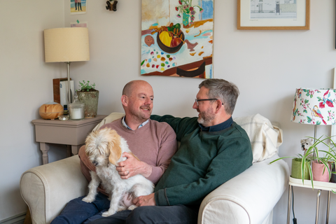 Two men sit inside their home on a white couch with a white dog