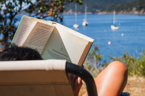 person reads book while sitting in chair looking out at lake and sailboats