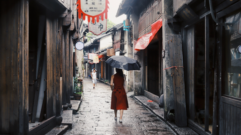 Woman walks in an alley in the rain wearing a red dress and carrying a black umbrella