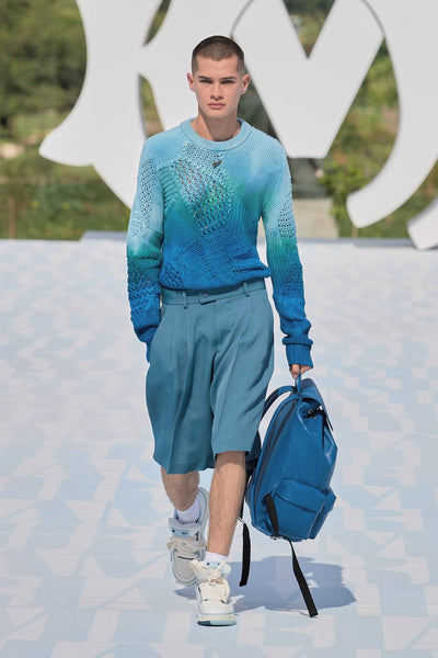 Model wears a blue sweatshirt, blue tailored shorts and high-top sneakers at AMIRI's SS23 presentation for Paris Fashion Week