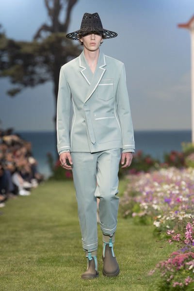 Model wears a pale blue tailored suit with a woven sunhat and rubber rain boots at Dior's SS23 presentation for Paris Fashion Week