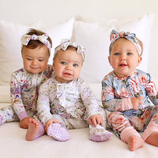 three infants sitting on bed together wearing floral print onesies and headwraps