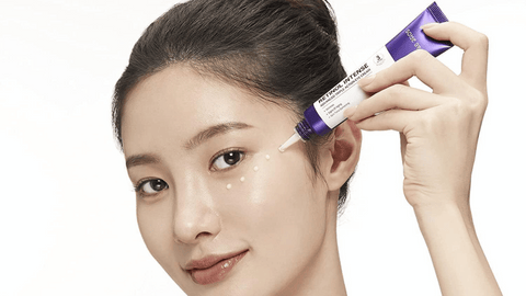 A woman applies small drops of a skincare product from a purple tube to her face. She looks at the camera with a soft smile and natural makeup. The background is clean and neutral.