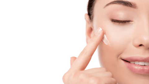 A woman is applying cream to her cheekbone. Her eyes are closed and her facial expression is relaxed. The background is white and clean, highlighting her skin and the cream she is applying.