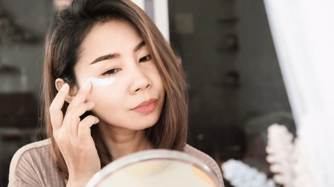 A woman applies a white face cream under her eye. She looks reflective, looking at herself in a mirror. Her hair is loose and the background is blurred, giving focus to her skincare routine.