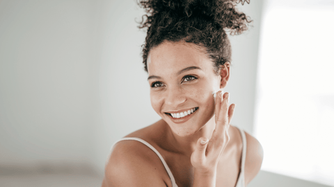 A smiling woman with her hair up is applying cream to her face. She looks happy and content, with a natural beauty highlighted in a bright and airy room.