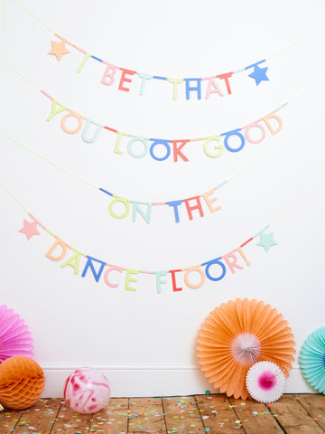 Neon letters that spell out "i bet that you look good on the dance floor" in 4 seperate swags. There is an orange paper honeycomb decocration, a pink paper fan, an orange paper fan and a turquoise paper fan on the floor underneath.