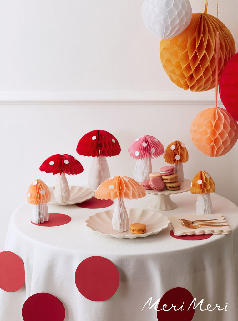 Round table set with cream plates and honeycomb toadstool decorations in red, pink, coral and yellow. There are honeycomb balls hanging from the ceiling on the right hand side of the image.