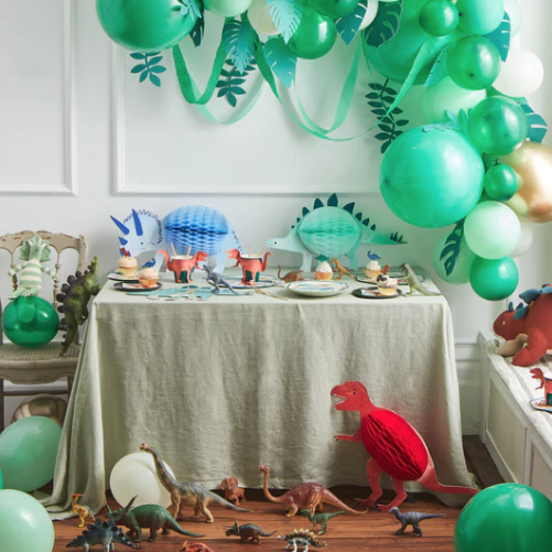 Meri Meri dinosaur party decorations on a table with a green balloon arch above it. There are paper honeycomb dinosaur decorations beside and on top of the table.